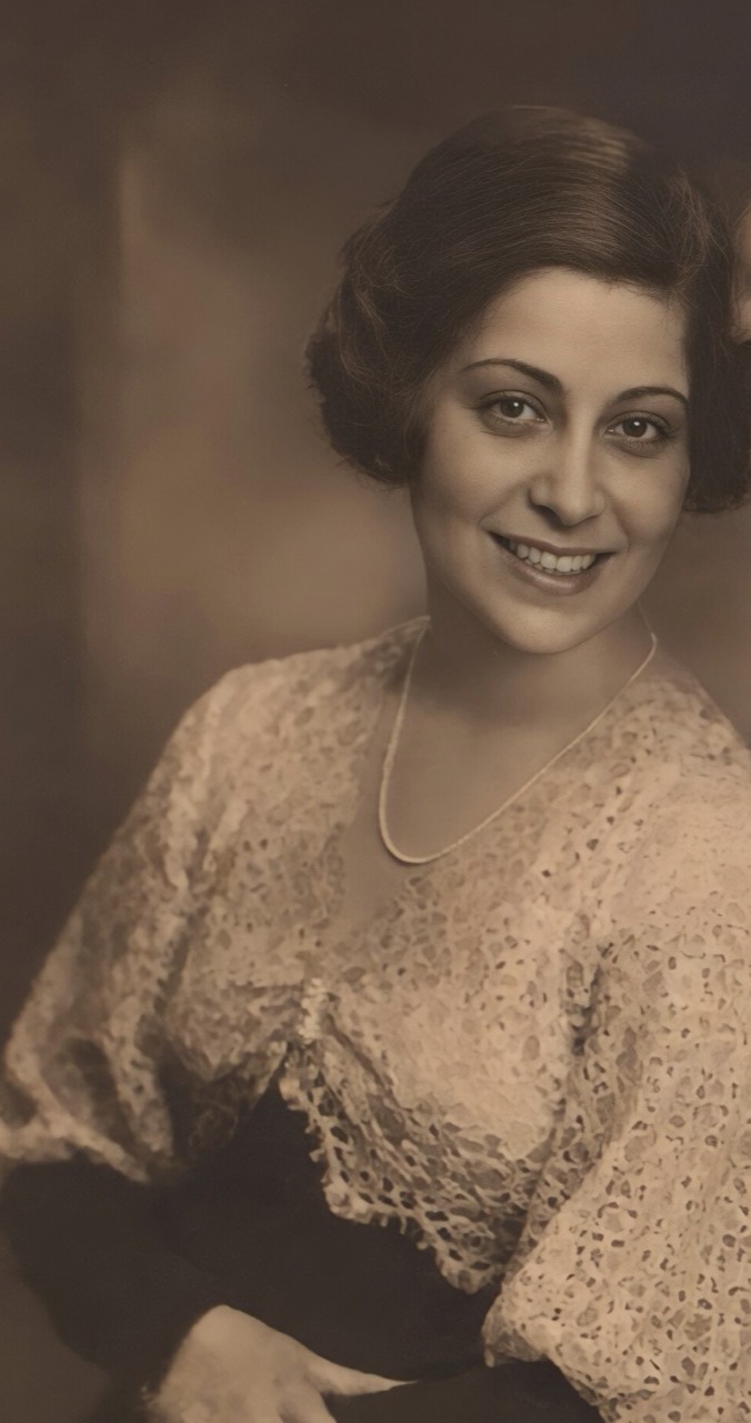 Full-length seated studio portrait of Ruth Helene Reichenbach as a young adult, smiling at the camera in a lace garment and necklace